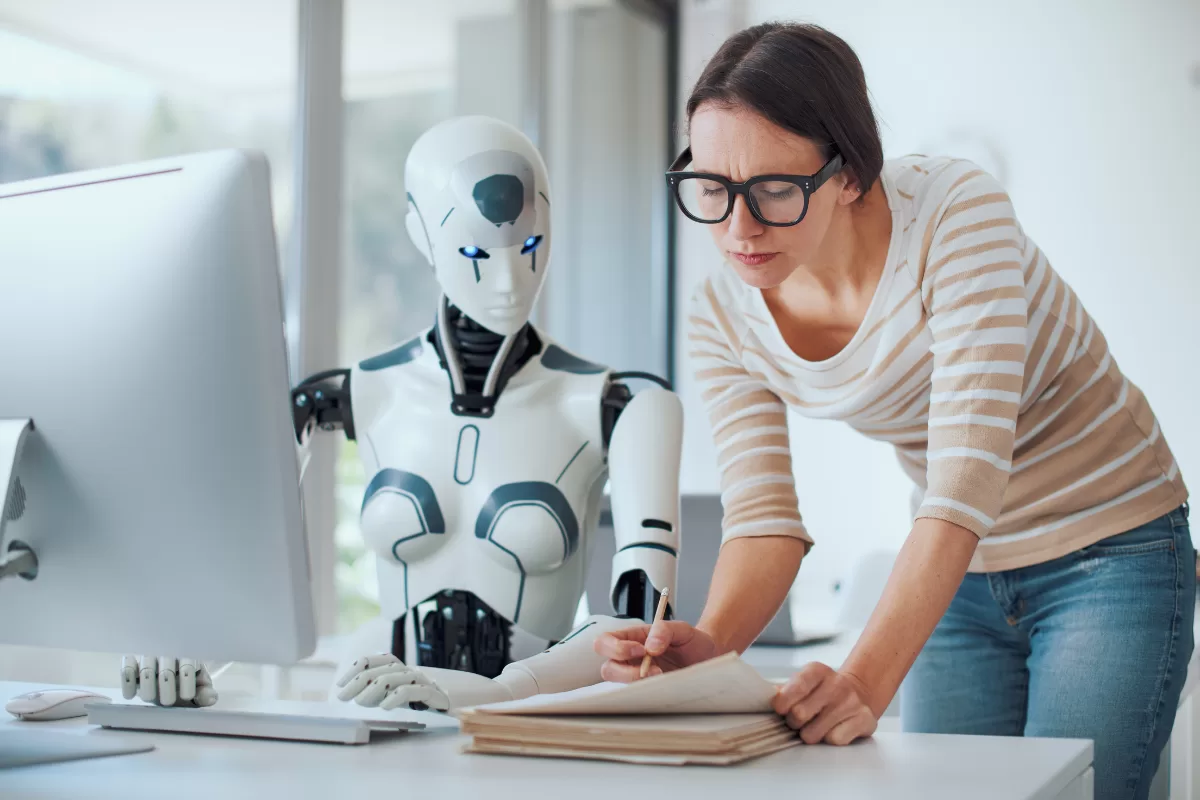 A humanoid robot sits beside a woman in a modern office, both focused on documents and a computer screen, illustrating human-AI collaboration in the workplace, representing AI agents