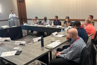 TestBooster's CEO standing and presenting at a conference room podium while several participants sit around U-shaped tables, listening and taking notes during a professional meeting, representing TestBooster at the Midsize Enterprise Summit
