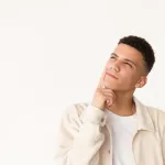 Um jovem pensativo de cabelo escuro e curto, vestindo uma jaqueta bege clara, olha para cima e ligeiramente para o lado com uma mão apoiada no queixo contra um fundo branco.