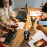 Três profissionais de tecnologia trabalhando colaborativamente em mesa compartilhada com laptops, tablet e anotações durante sessão de bug bash