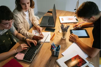 Três profissionais de tecnologia trabalhando colaborativamente em mesa compartilhada com laptops, tablet e anotações durante sessão de bug bash
